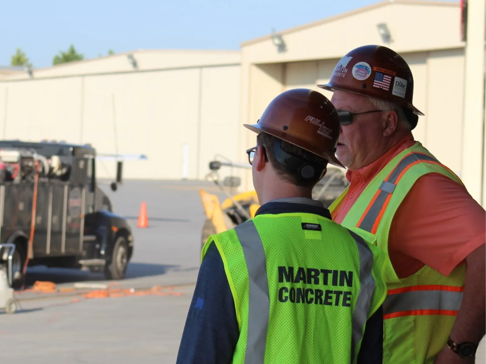 Two construction workers with safety vests and helmets discussing on site near equipment and buildings.