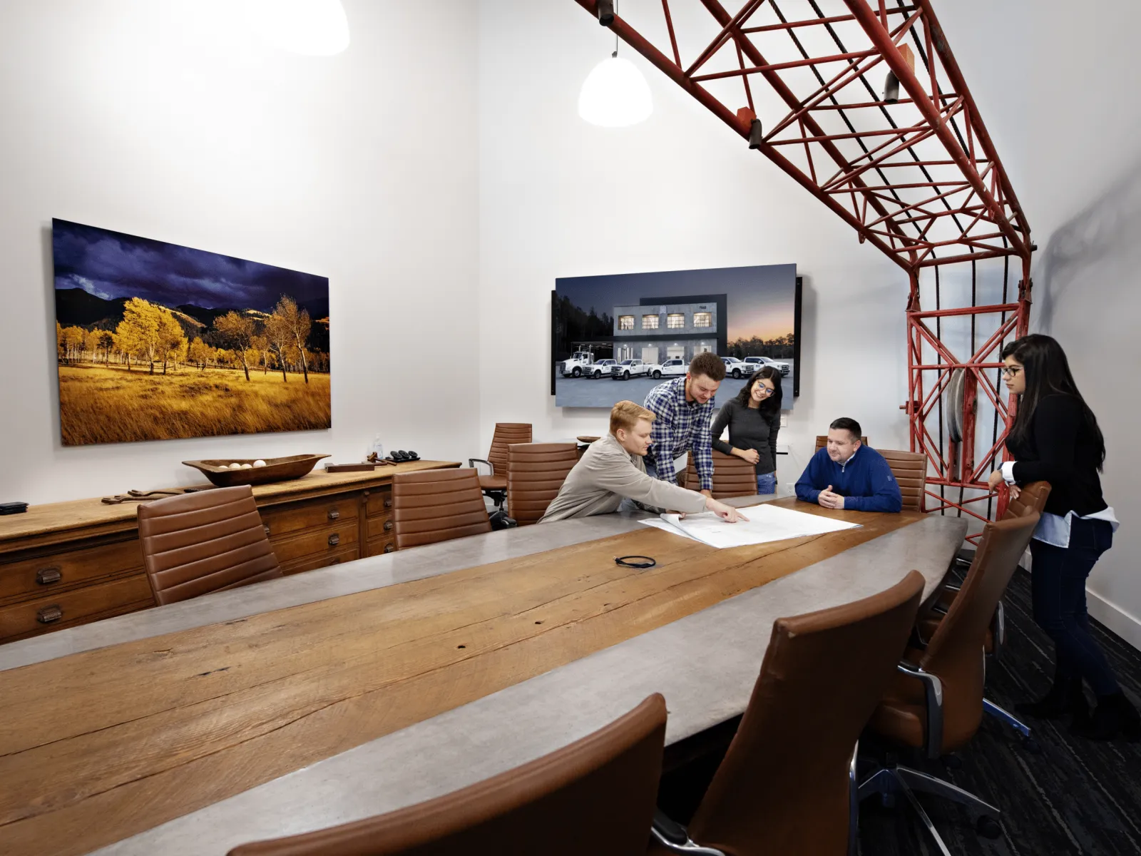 Team of five collaborating over architectural plans in modern conference room with artwork and industrial ceiling detail