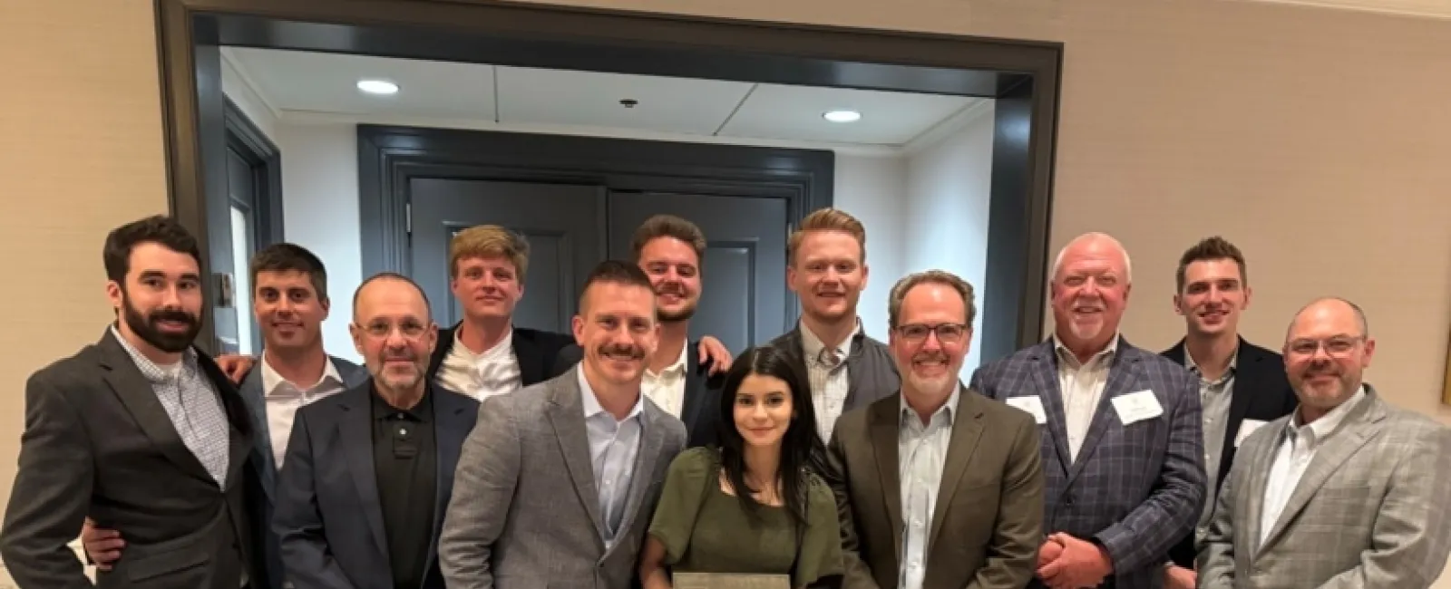 Group of eleven people in business casual attire posing indoors, woman in front holding an award plaque.