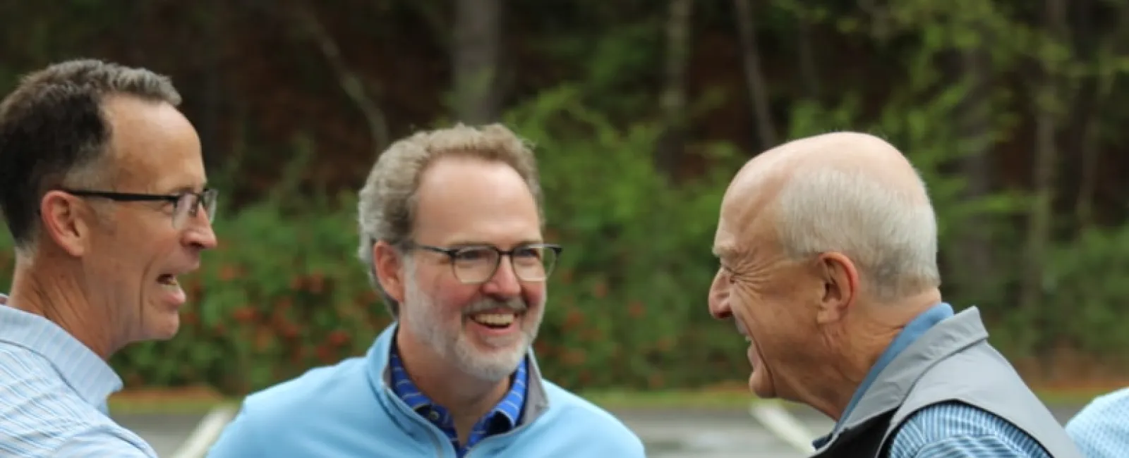 Three men outdoors smiling and shaking hands during a friendly meeting in a parking lot with greenery background