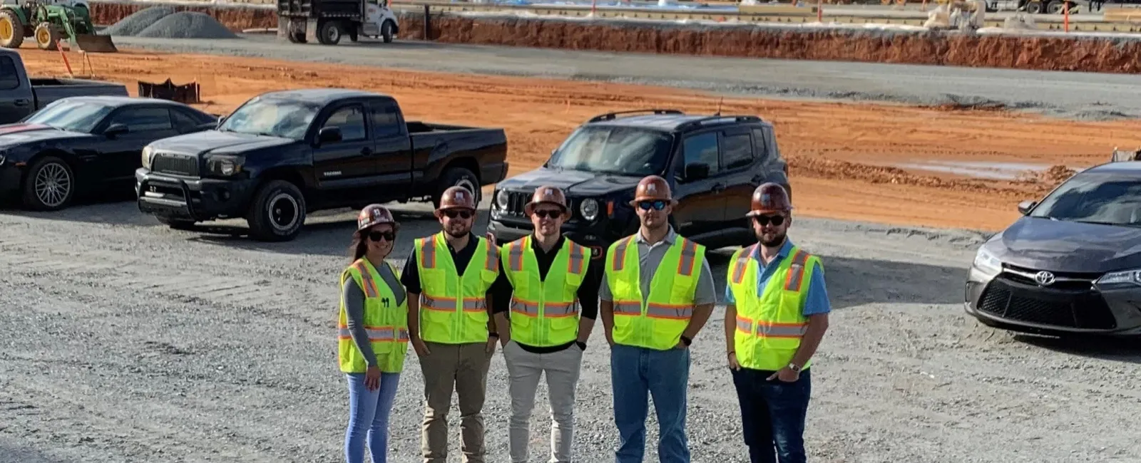 Five construction workers in safety vests and helmets standing on a gravel site with trucks and equipment nearby.