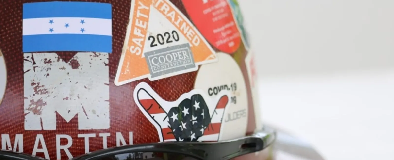 Brown construction helmet with various stickers and black safety glasses resting on it on a light background.