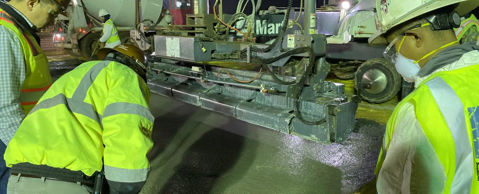 Construction workers in safety gear operating machinery pouring and smoothing concrete at night on a road project.