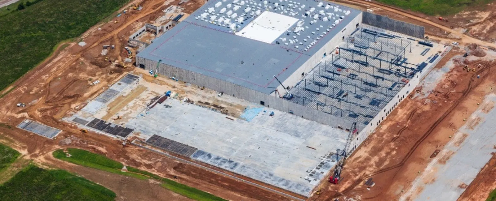 Aerial view of a large industrial building under construction surrounded by green fields and dirt roads.
