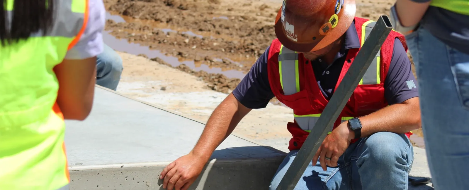 Construction worker in safety gear inspecting freshly poured concrete curb at an outdoor building site.