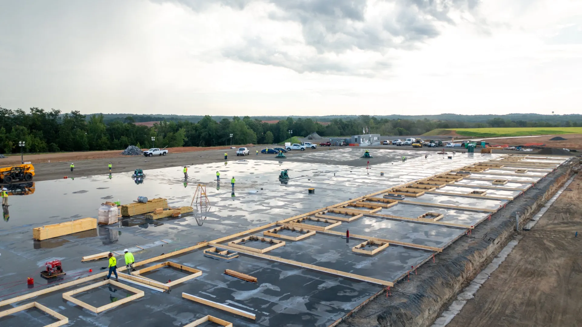 Construction site with wet concrete slab and workers preparing wooden formwork under cloudy sky.