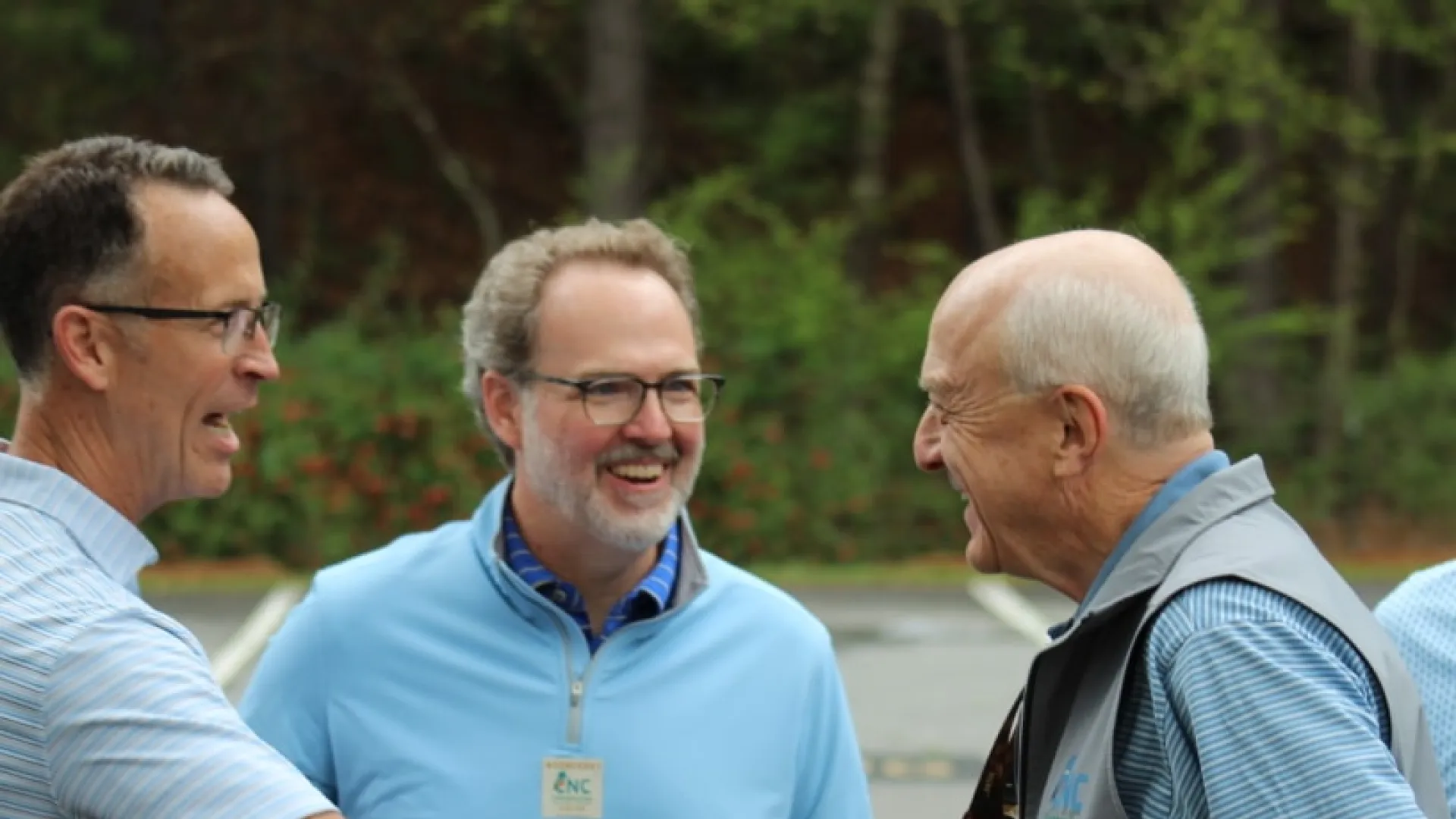 Three men outdoors smiling and shaking hands during a friendly meeting in a parking lot with greenery background