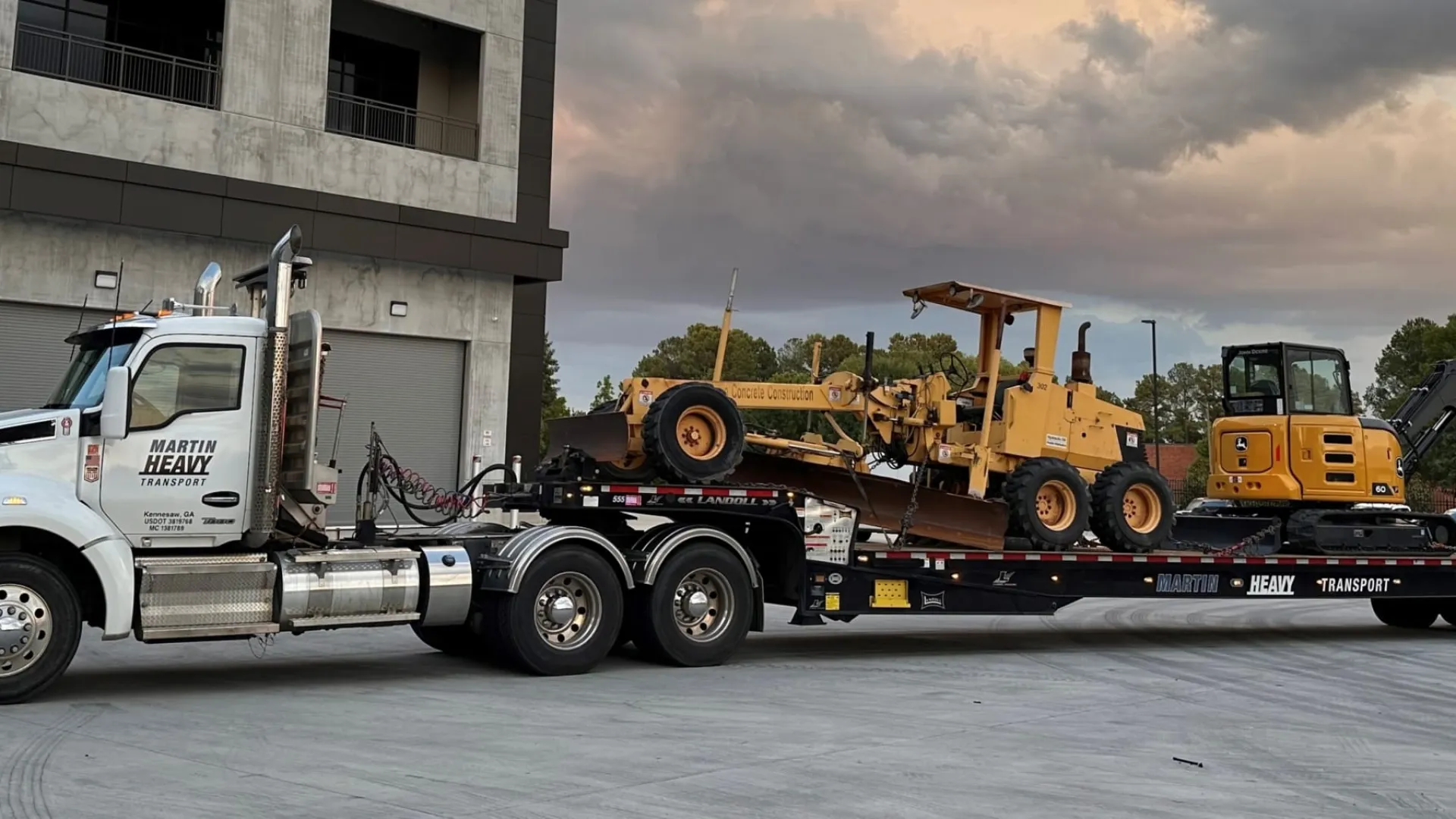 White heavy-duty truck transporting large construction machinery on flatbed trailer at industrial site.