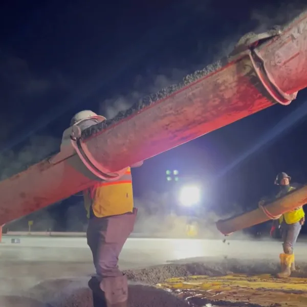 Workers pouring concrete at a nighttime construction site with bright floodlights and large hoses.