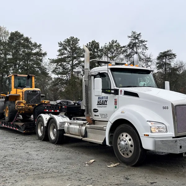 White heavy-duty truck carrying yellow construction equipment on a flatbed trailer in a forested area.