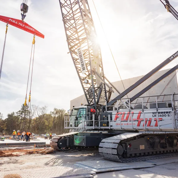 Large crawler crane lifting concrete slabs at a construction site under clear sky with workers in safety gear.