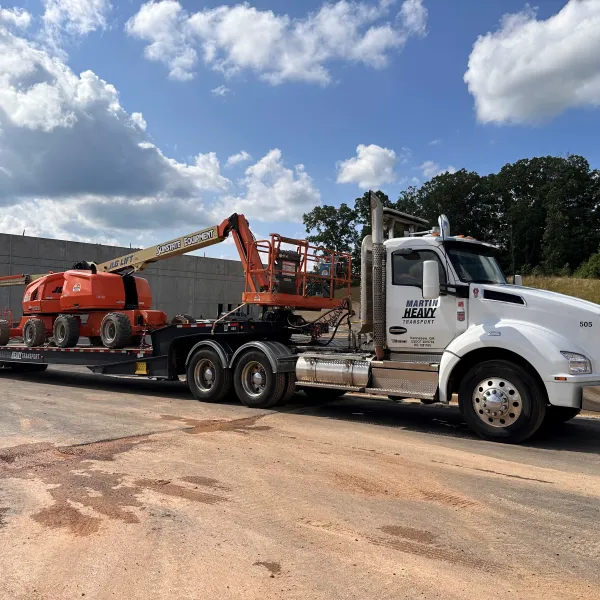 White heavy transport truck carrying orange construction lift equipment on a flatbed trailer on a sunny day.