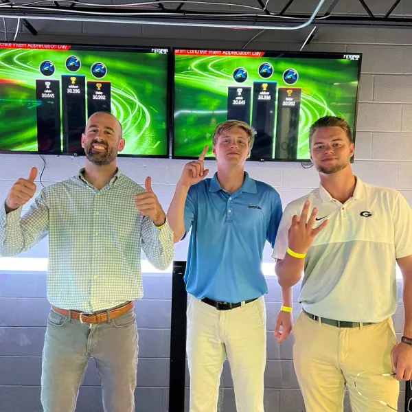 Three men in casual shirts posing with thumbs up and number signs in front of two monitors displaying scores.