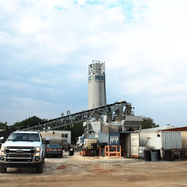 Concrete plant with mixing tower, conveyor belt, construction trucks, and vehicles under cloudy sky.