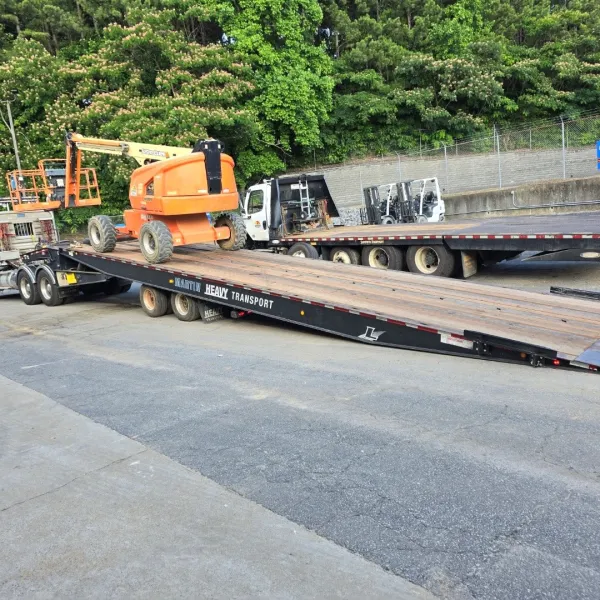 Orange boom lift being loaded onto a heavy transport flatbed trailer in a parking lot with trees in the background.