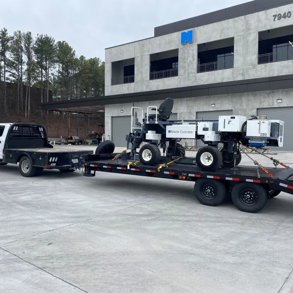Flatbed trailer carrying Martin Concrete power buggy attached to white pickup truck outside industrial building.