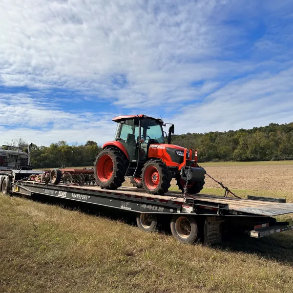 Red tractor loaded on a flatbed trailer attached to a semi truck in a rural field under a partly cloudy sky.
