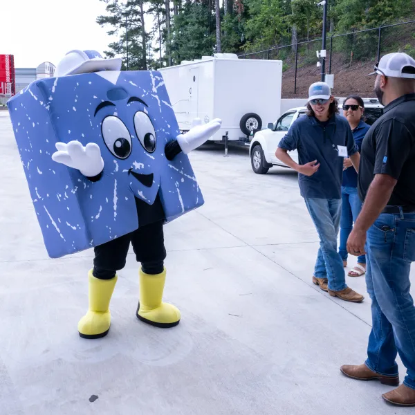 Person in blue block costume with yellow boots interacting with three people outside near parked vehicles.