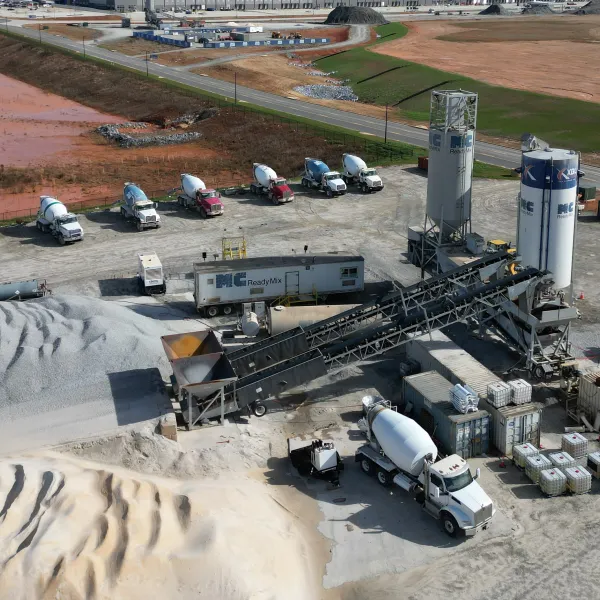 Aerial view of a concrete batching plant with cement trucks, silos, conveyor belts, and raw materials piles.