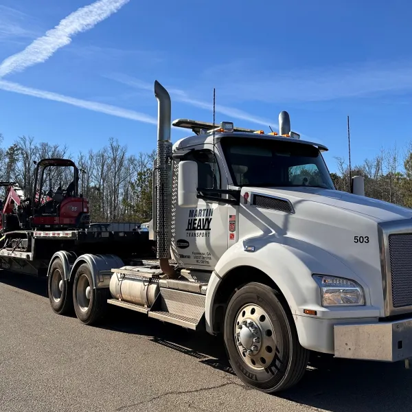 White heavy-duty Kenworth truck carrying red compact excavator on flatbed trailer on clear day