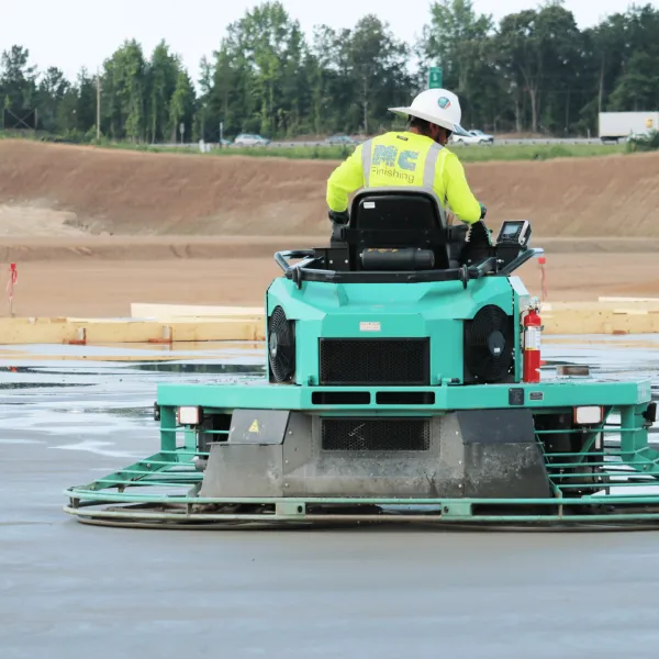 Construction worker operating a green power trowel to smooth wet concrete on a large outdoor slab.