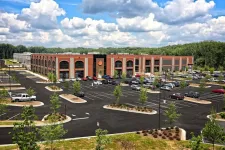 Thumbnail for Modern red brick commercial building with large parking lot surrounded by trees under a partly cloudy sky