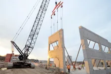 Thumbnail for Crane lifting large concrete wall panel at construction site with workers and steel supports during sunset.