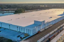 Thumbnail for A large warehouse with railcars on adjacent train tracks and a pond visible in the background at sunrise.