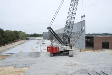 Thumbnail for Crane lifting a large concrete wall panel at a construction site with surrounding unfinished structures and trees.