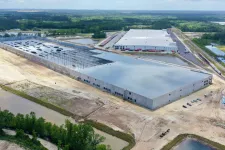 Thumbnail for Aerial view of large industrial warehouse buildings surrounded by construction and water bodies under a partly cloudy sky.