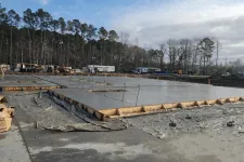 Thumbnail for Freshly poured concrete slab foundation at a construction site surrounded by trees and equipment under cloudy sky
