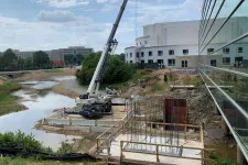 Thumbnail for Crane and construction site by a river with steel reinforcements and workers near a large modern building.