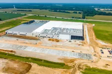 Thumbnail for Aerial view of a large industrial warehouse under construction surrounded by fields and a dirt parking area.