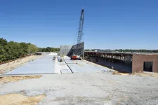 Thumbnail for Construction site with concrete slabs, crane lifting wall panels, and partially built structures under clear blue sky.
