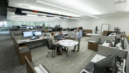 Modern office with open workstations and three employees discussing plans at a round table