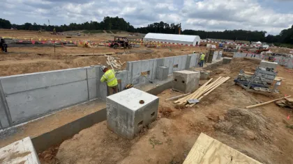 Construction site with workers installing concrete walls and blocks under a cloudy sky on bare earth ground.