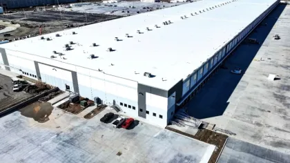 Aerial view of a large industrial warehouse with white roof surrounded by concrete pavement and few parked vehicles.