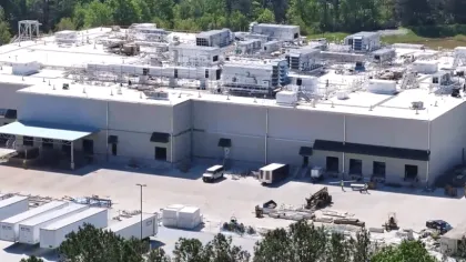 A large industrial warehouse surrounded by trees with multiple rooftop HVAC units and trucks outside.