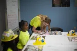 Instructor helps children in yellow shirts and hard hats during a hands-on learning activity at a table.
