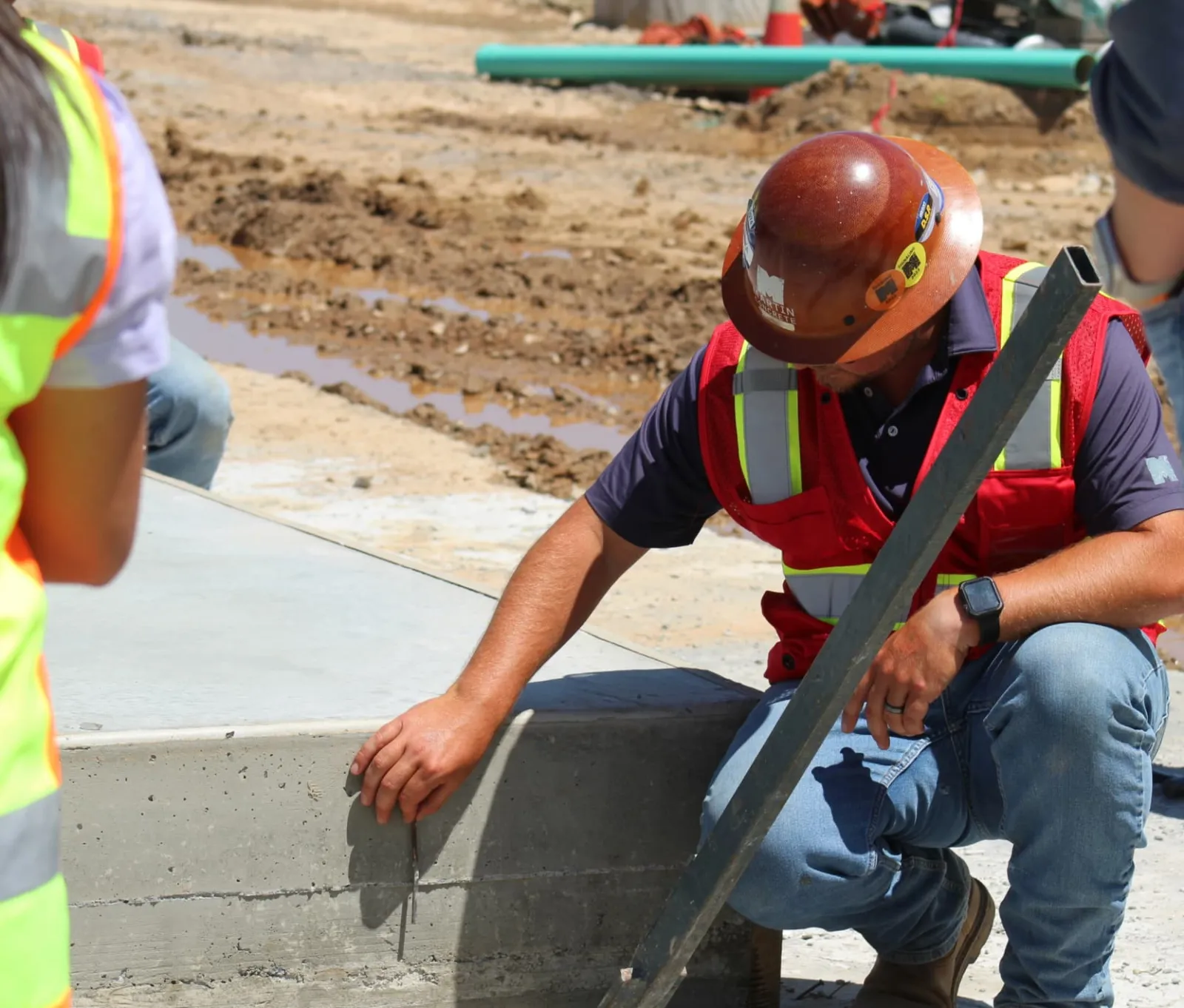 Construction worker in safety gear inspecting freshly poured concrete curb at an outdoor site under sunny conditions