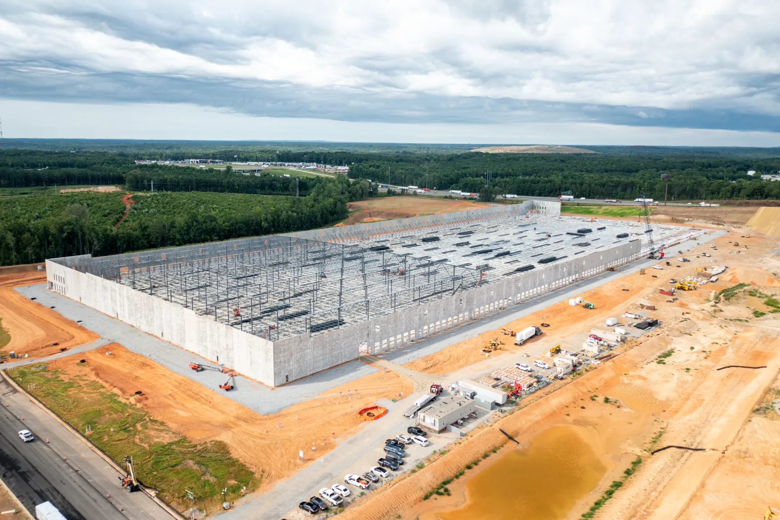 A large warehouse building under construction surrounded by dirt and construction vehicles under cloudy skies.
