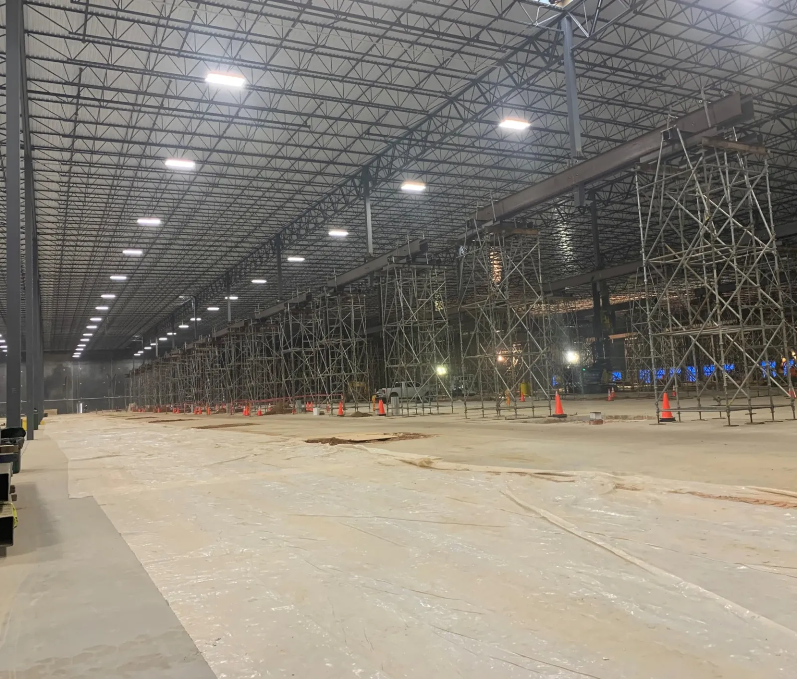 Large industrial warehouse interior under construction with metal scaffolding and overhead lighting.
