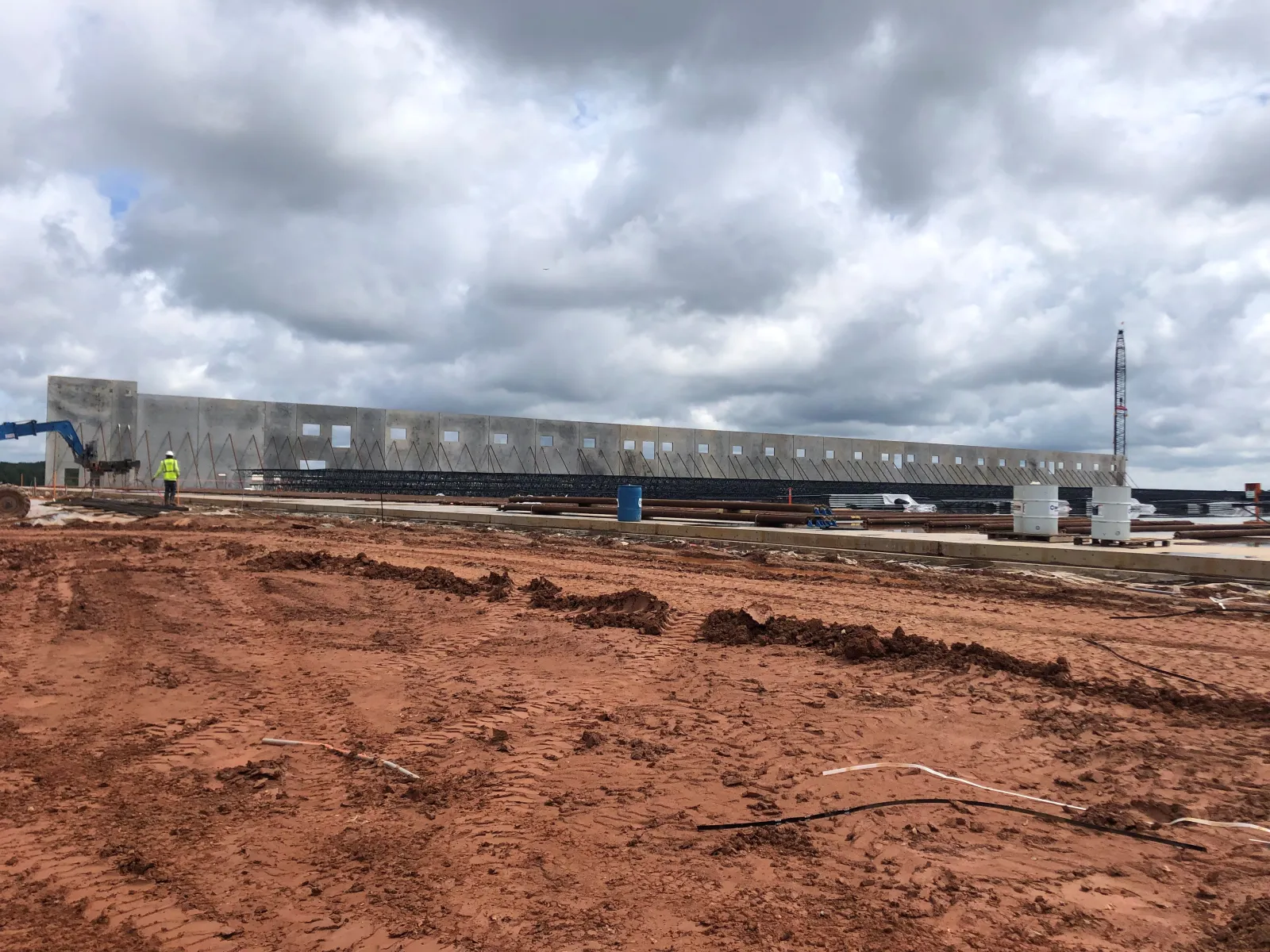 Construction site with large concrete wall panels being erected under cloudy sky on red dirt ground.