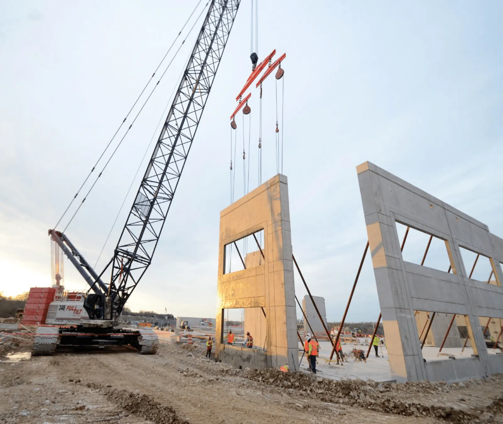Crane lifting large concrete wall panel at construction site with workers and steel supports during sunset.