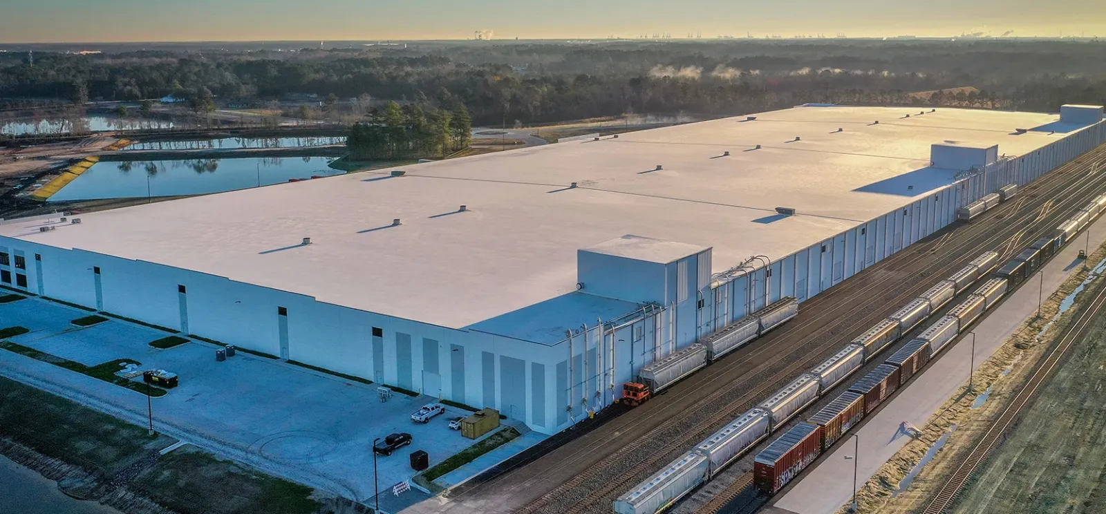 A large warehouse with railcars on adjacent train tracks and a pond visible in the background at sunrise.