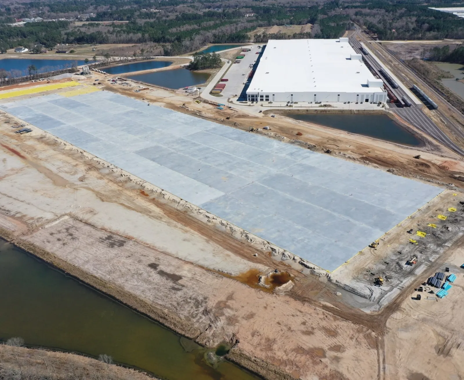 Aerial view of large warehouse construction site with concrete foundation and surrounding water bodies in a rural area.