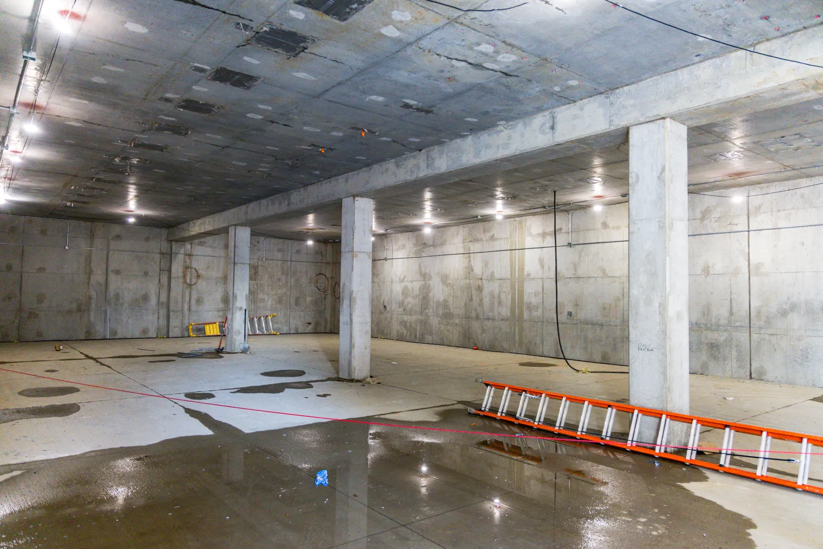 Empty concrete basement with support pillars, scattered water puddles, and orange ladder on the floor under ceiling lights.