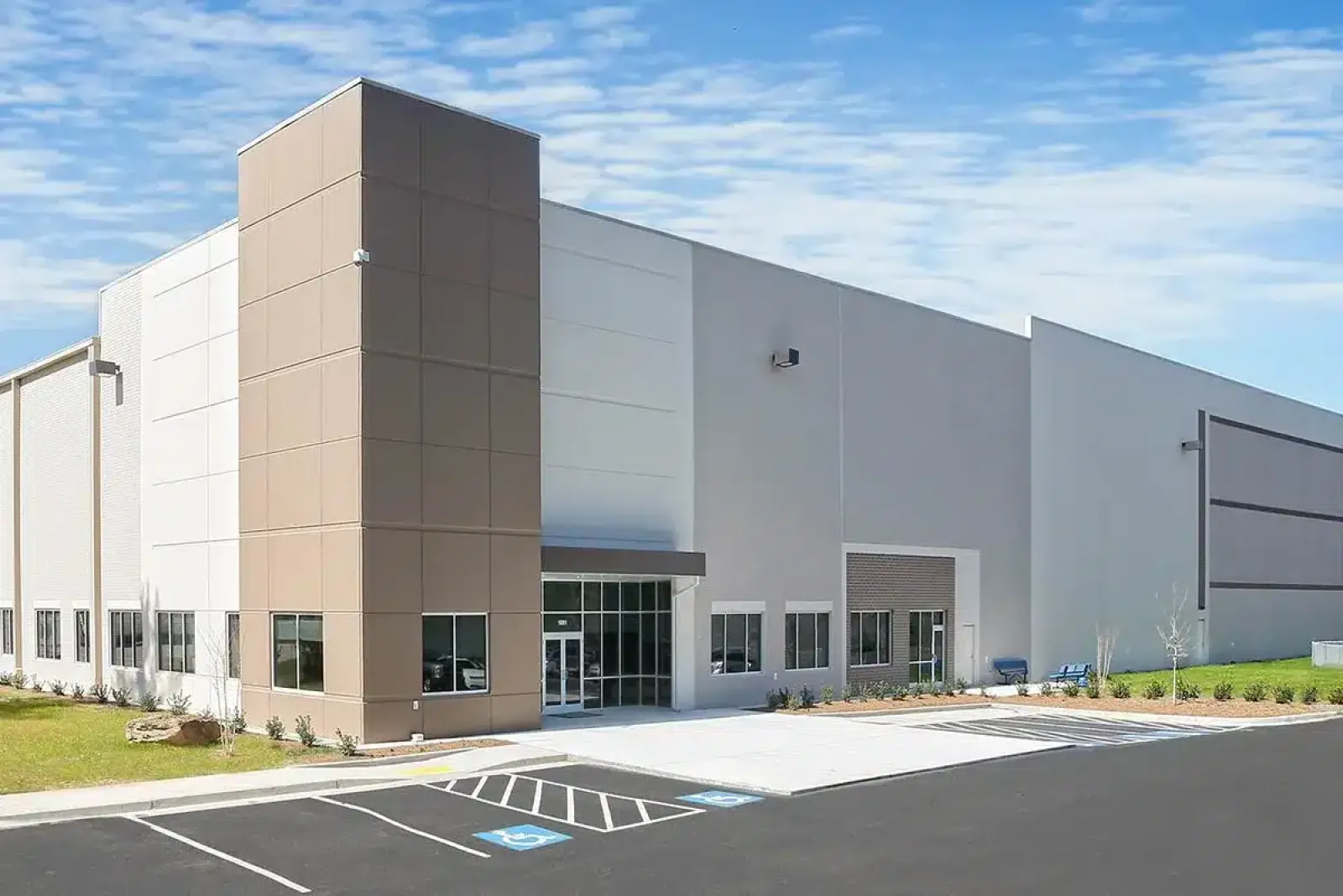 Modern industrial warehouse building with glass entrance and parking spaces under blue sky.