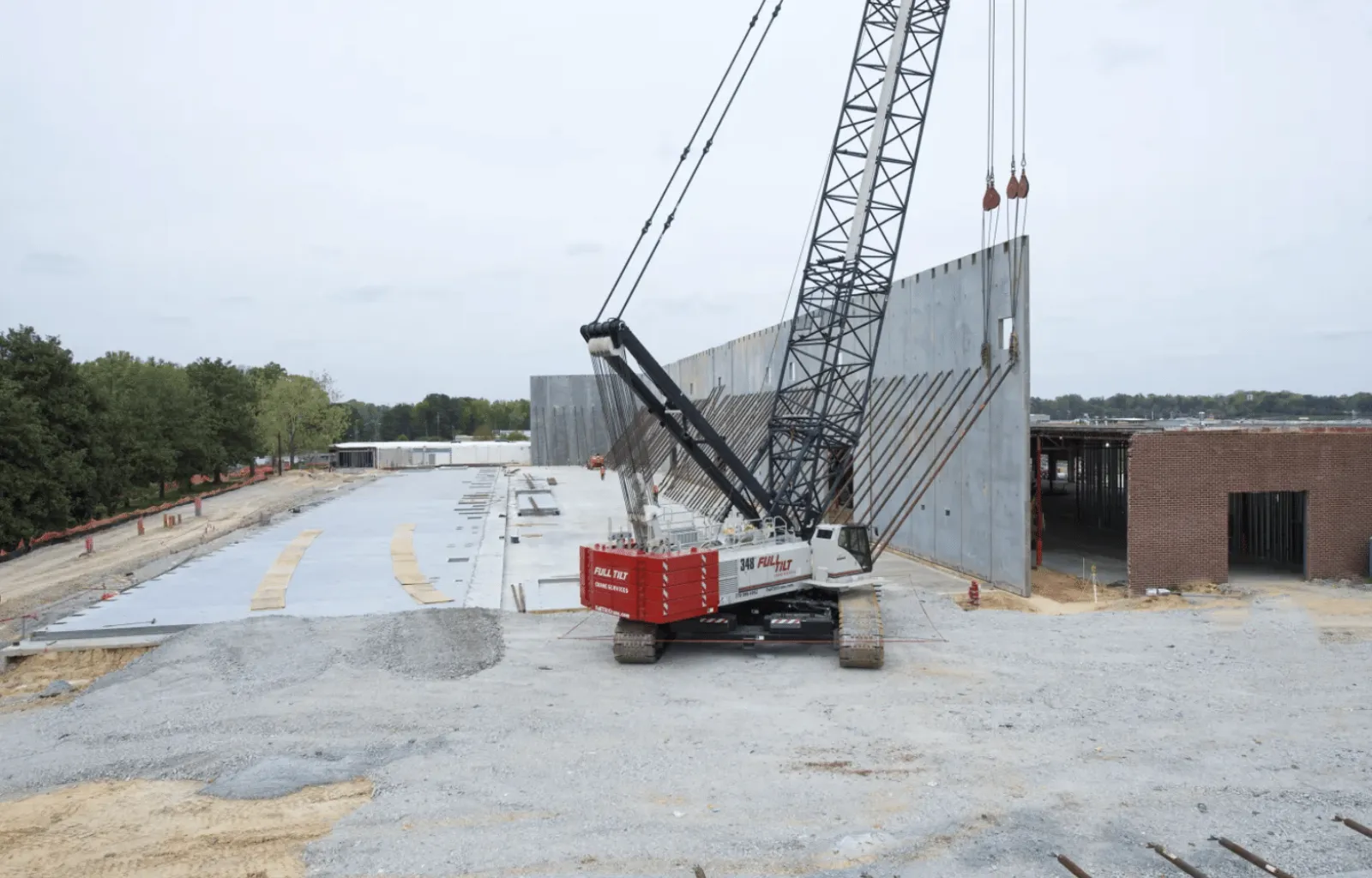Crane lifting a large concrete wall panel at a construction site with surrounding unfinished structures and trees.