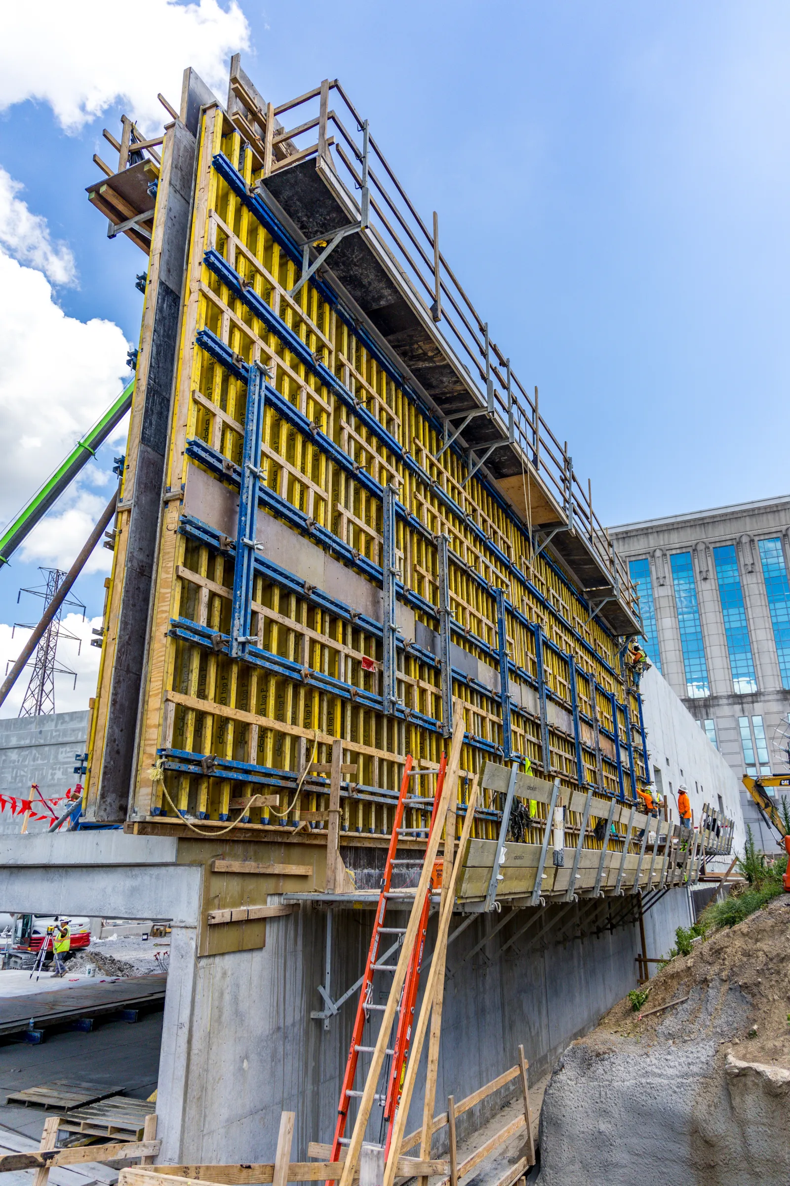 Large vertical concrete formwork with ladders and scaffolding at an urban construction site on a clear day.